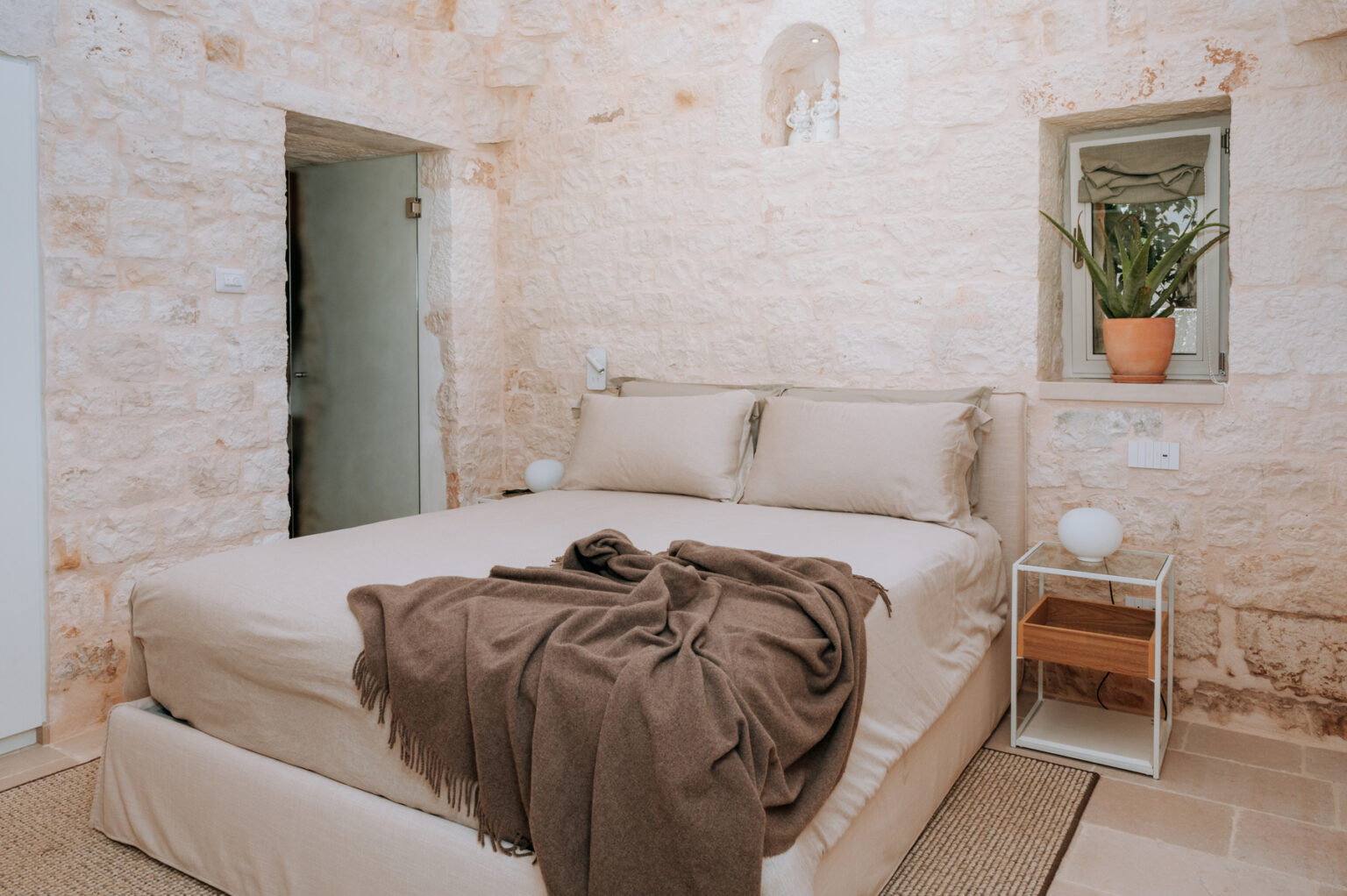 Cozy bedroom featuring a comfortable bed with beige linens, a brown throw blanket, and a small bedside table. The rustic stone walls and natural light create a warm and inviting atmosphere, enhanced by a potted plant near the window. | Only on SopranoVillas