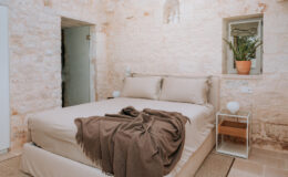 Cozy bedroom featuring a comfortable bed with beige linens, a brown throw blanket, and a small bedside table. The rustic stone walls and natural light create a warm and inviting atmosphere, enhanced by a potted plant near the window.