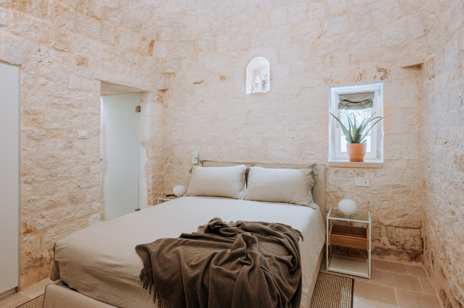 Cozy bedroom featuring a stone wall design, a comfortable bed with neutral bedding, a decorative throw, a minimalist nightstand, and a potted plant by the window, creating a serene and inviting atmosphere. | Only on SopranoVillas
