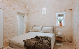 Cozy bedroom featuring a stone wall design, a comfortable bed with neutral bedding, a decorative throw, a minimalist nightstand, and a potted plant by the window, creating a serene and inviting atmosphere.