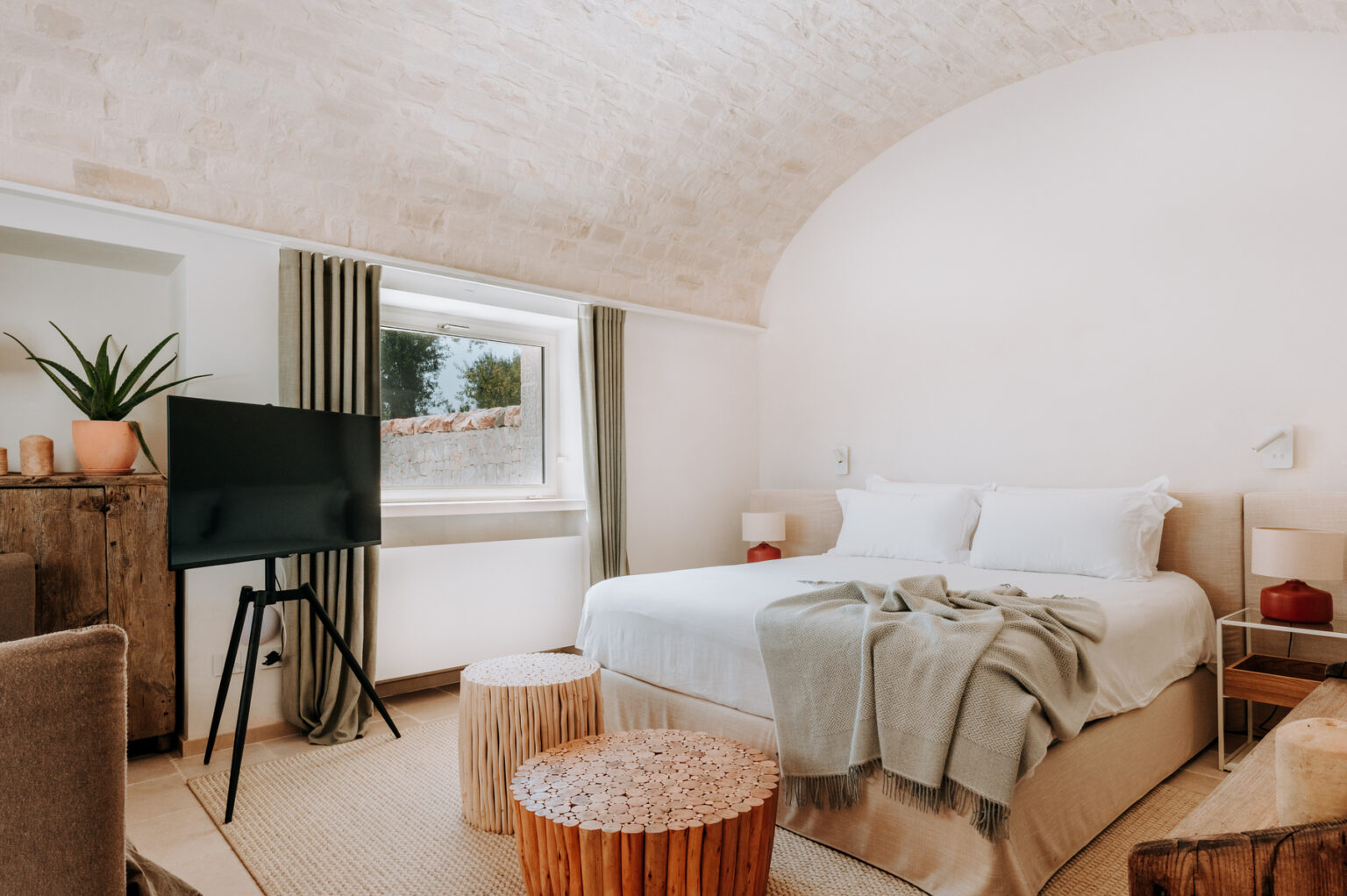 Cozy and modern bedroom featuring a comfortable bed with soft linens, stylish decor, a flat-screen TV, and natural light from a window. The room includes textured seating and decorative elements like a plant and woven stools, creating a warm and inviting atmosphere. | Only on SopranoVillas