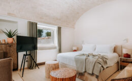 Cozy and modern bedroom featuring a comfortable bed with soft linens, stylish decor, a flat-screen TV, and natural light from a window. The room includes textured seating and decorative elements like a plant and woven stools, creating a warm and inviting atmosphere.