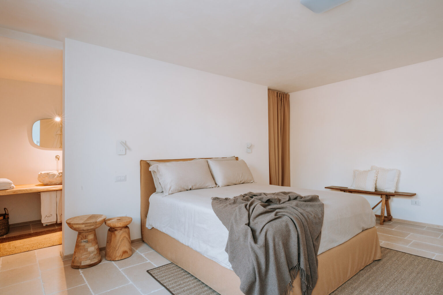 Modern bedroom featuring a comfortable bed with neutral bedding, minimalist decor, wooden furniture, and a cozy atmosphere. The space includes a stylish bathroom area visible in the background, enhancing the room's inviting aesthetic. | Only on SopranoVillas