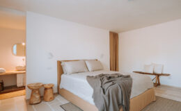 Modern bedroom featuring a comfortable bed with neutral bedding, minimalist decor, wooden furniture, and a cozy atmosphere. The space includes a stylish bathroom area visible in the background, enhancing the room's inviting aesthetic.