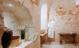 Cozy interior of a rustic stone bathroom featuring natural light, a wooden table, decorative plants, and minimalist decor, creating a serene atmosphere.