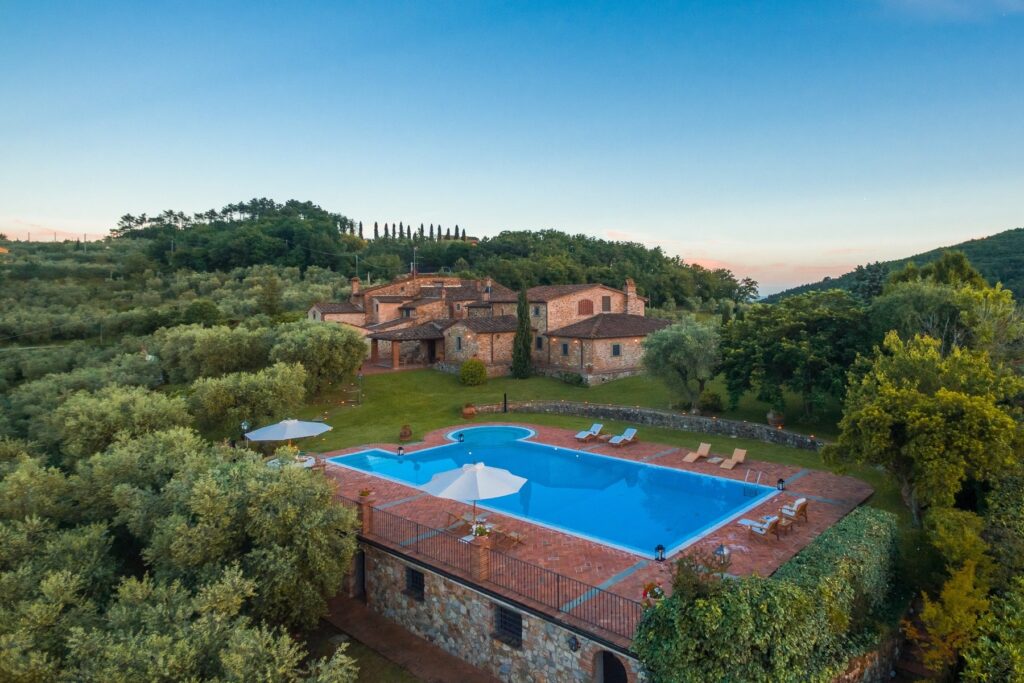 Aerial view of a luxurious villa with a blue swimming pool surrounded by lush greenery and rolling hills at sunset. Ideal for vacation rentals in a serene countryside setting. | Only on SopranoVillas