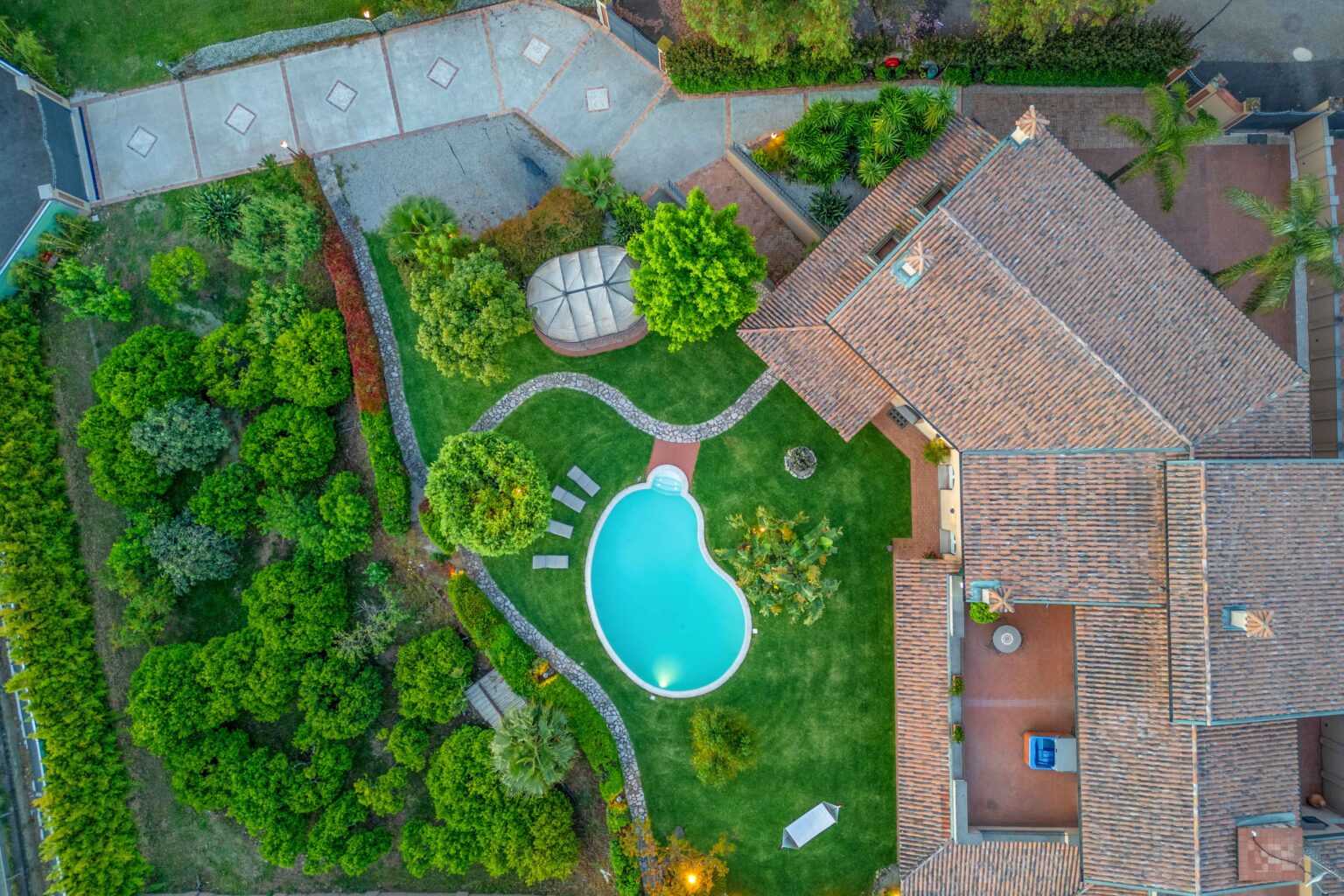 Aerial view of a luxurious residential property featuring a landscaped garden, a swimming pool in an organic shape, and a cozy gazebo. The well-maintained lawn is surrounded by lush greenery and pathways, showcasing a harmonious blend of nature and modern architecture. Ideal for outdoor relaxation and entertaining. | Only on SopranoVillas