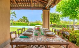 Outdoor dining area featuring a beautifully set wooden table with decorative plates and glassware, surrounded by lush greenery and a swimming pool in the background, ideal for al fresco meals.