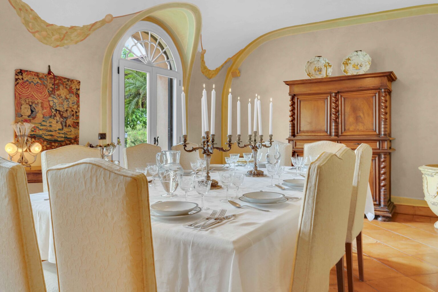 Elegant dining room featuring a beautifully set table with white tablecloth, fine china, and crystal glassware. A candelabra with white candles serves as a centerpiece, complemented by a wooden cabinet and decorative artwork. Large windows allow natural light and a view of greenery outside, enhancing the inviting atmosphere. | Only on SopranoVillas