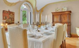 Elegant dining room featuring a beautifully set table with white tablecloth, fine china, and crystal glassware. A candelabra with white candles serves as a centerpiece, complemented by a wooden cabinet and decorative artwork. Large windows allow natural light and a view of greenery outside, enhancing the inviting atmosphere.