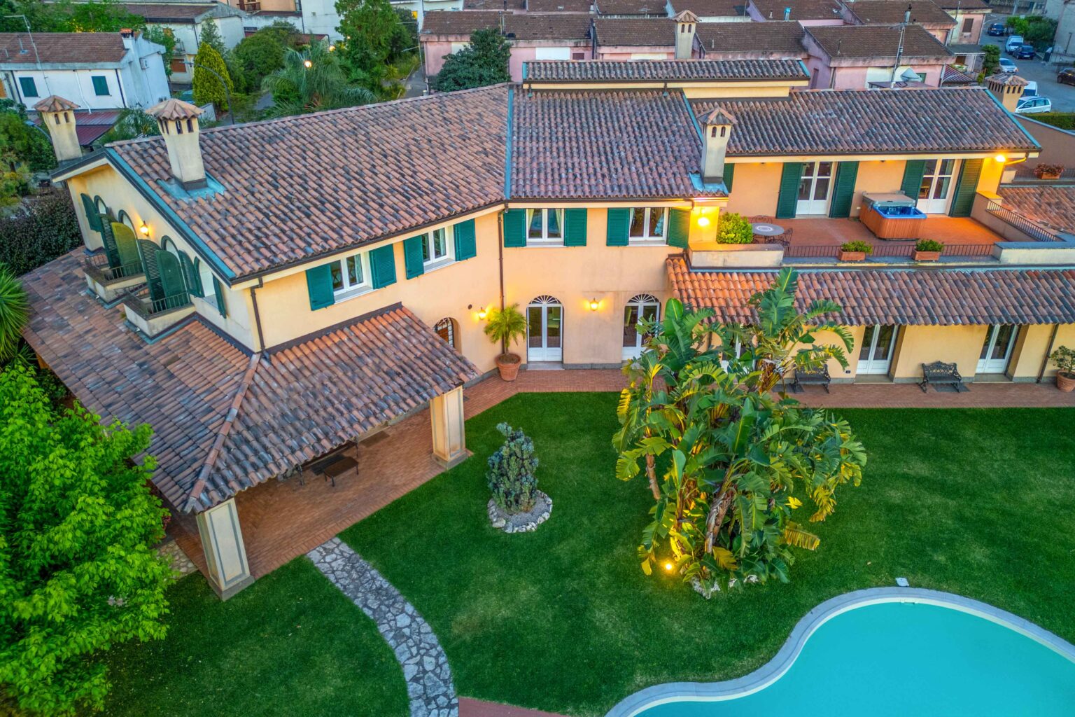Aerial view of a charming villa featuring a tiled roof, green shutters, and a lush garden with a swimming pool. The property includes a spacious outdoor area with decorative plants and a stone pathway leading to the entrance. | Only on SopranoVillas