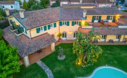Aerial view of a charming villa featuring a tiled roof, green shutters, and a lush garden with a swimming pool. The property includes a spacious outdoor area with decorative plants and a stone pathway leading to the entrance.