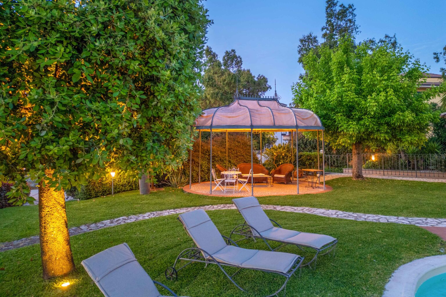 A serene garden setting featuring a gazebo surrounded by lush greenery, with comfortable seating and a small table. Lounge chairs are arranged on the manicured lawn, creating a perfect outdoor relaxation space. Soft lighting enhances the tranquil atmosphere during dusk. | Only on SopranoVillas