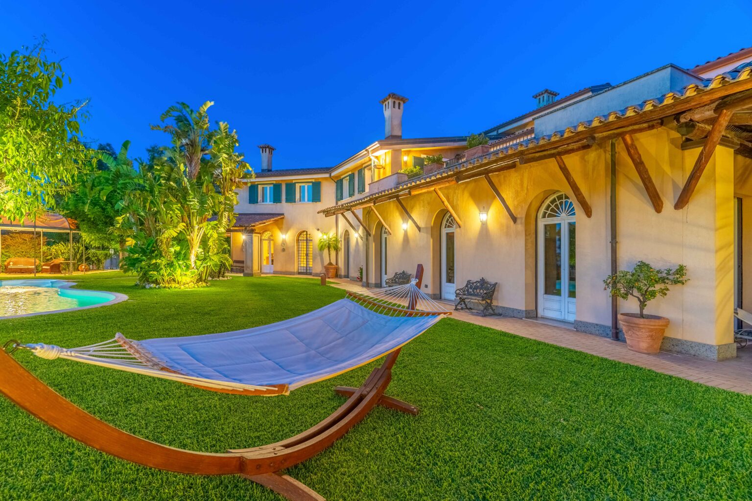 Luxurious villa with a hammock in a lush garden, featuring a swimming pool and beautifully landscaped greenery, illuminated at dusk. | Only on SopranoVillas