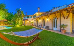 Luxurious villa with a hammock in a lush garden, featuring a swimming pool and beautifully landscaped greenery, illuminated at dusk.