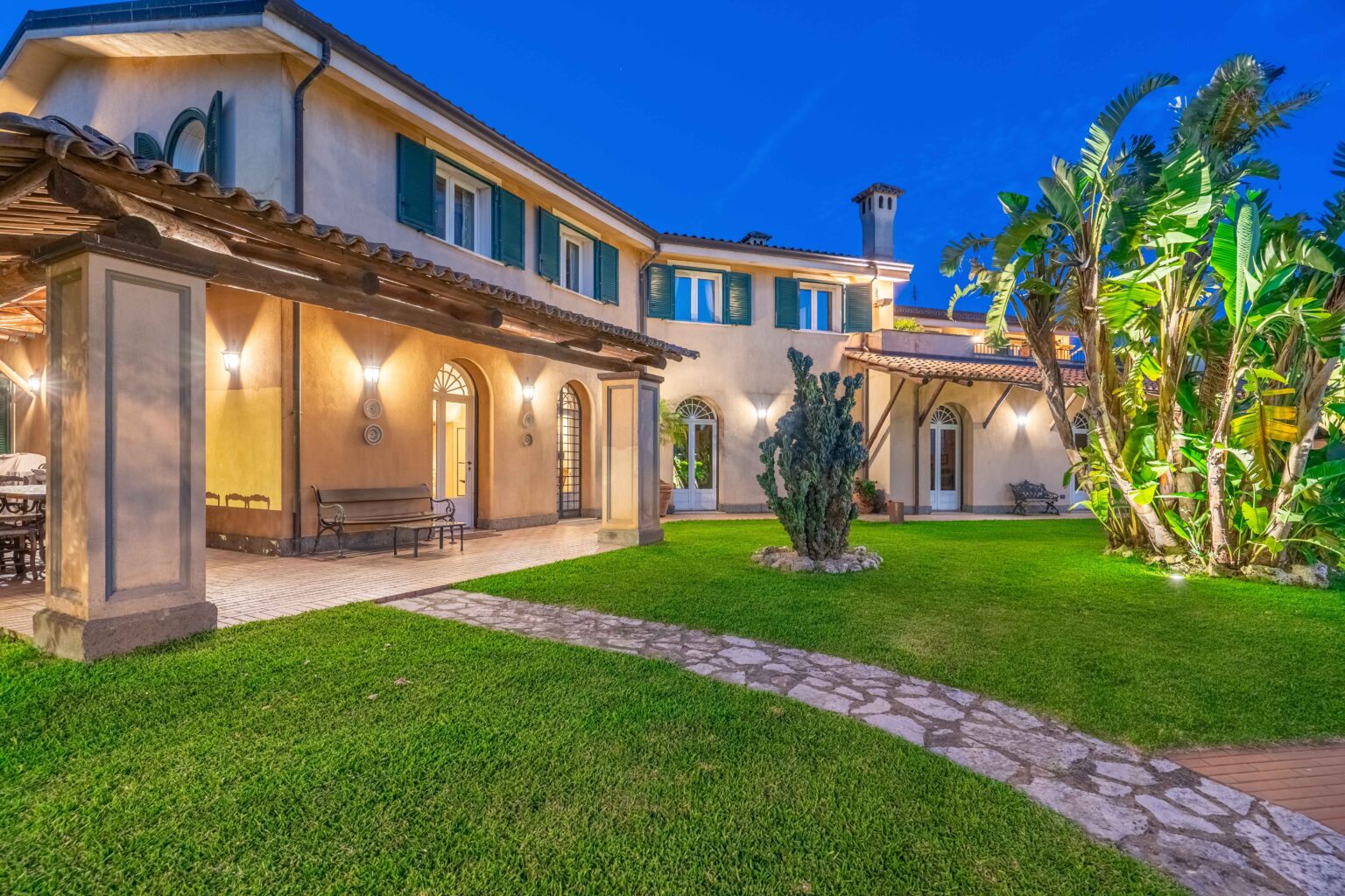Elegant villa exterior at dusk, showcasing a charming garden, stone pathway, and tropical plants. The architecture features arched windows and a tiled roof, creating a warm and inviting atmosphere. Perfect for outdoor gatherings and relaxation. | Only on SopranoVillas