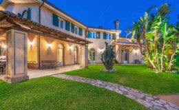 Elegant villa exterior at dusk, showcasing a charming garden, stone pathway, and tropical plants. The architecture features arched windows and a tiled roof, creating a warm and inviting atmosphere. Perfect for outdoor gatherings and relaxation.