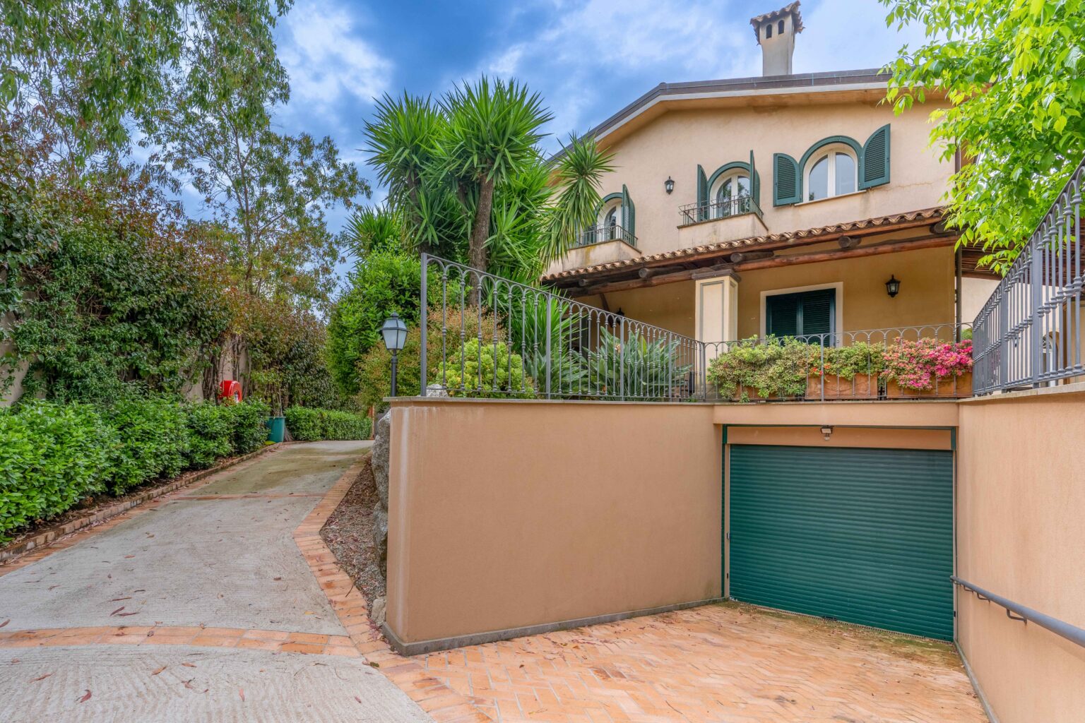 Charming residential property featuring a well-maintained driveway leading to a two-story house with green shutters, surrounded by lush greenery and decorative plants. The image showcases a garage entrance and a welcoming porch area, set against a partly cloudy sky. | Only on SopranoVillas
