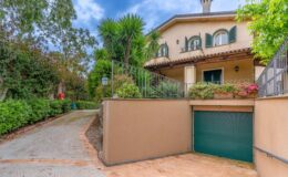 Charming residential property featuring a well-maintained driveway leading to a two-story house with green shutters, surrounded by lush greenery and decorative plants. The image showcases a garage entrance and a welcoming porch area, set against a partly cloudy sky.