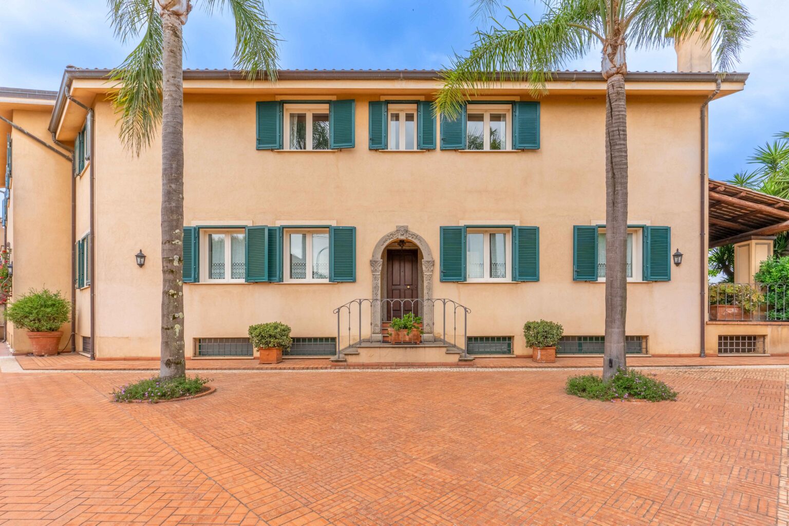 Two-story residential building with beige exterior, green shutters, and a welcoming entrance. The property features potted plants, palm trees, and a brick driveway, surrounded by a well-maintained landscape. Ideal for showcasing Mediterranean architecture and outdoor living. | Only on SopranoVillas