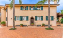 Two-story residential building with beige exterior, green shutters, and a welcoming entrance. The property features potted plants, palm trees, and a brick driveway, surrounded by a well-maintained landscape. Ideal for showcasing Mediterranean architecture and outdoor living.