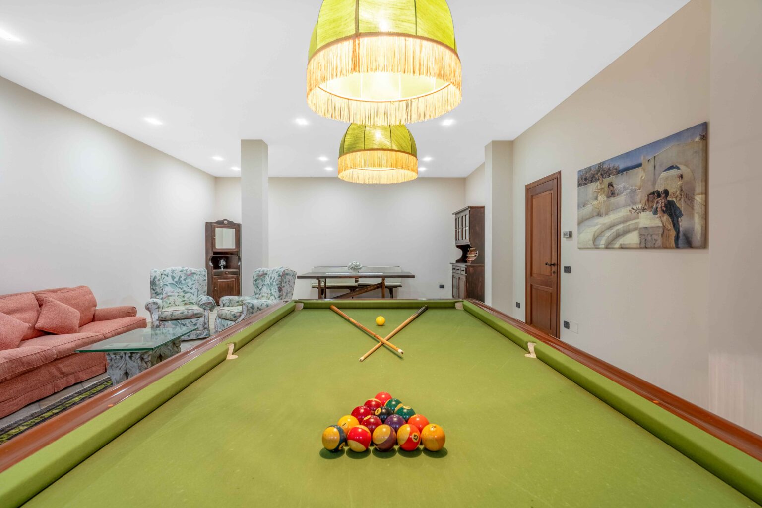 Interior view of a game room featuring a green pool table with a triangle of colored billiard balls, two cue sticks crossed on the table, and stylish overhead lights. The background includes cozy seating with floral patterns, a wooden sideboard, and a wall painting, creating a welcoming atmosphere for leisure activities. | Only on SopranoVillas