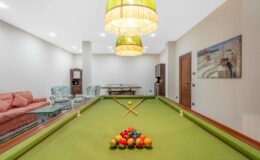 Interior view of a game room featuring a green pool table with a triangle of colored billiard balls, two cue sticks crossed on the table, and stylish overhead lights. The background includes cozy seating with floral patterns, a wooden sideboard, and a wall painting, creating a welcoming atmosphere for leisure activities.