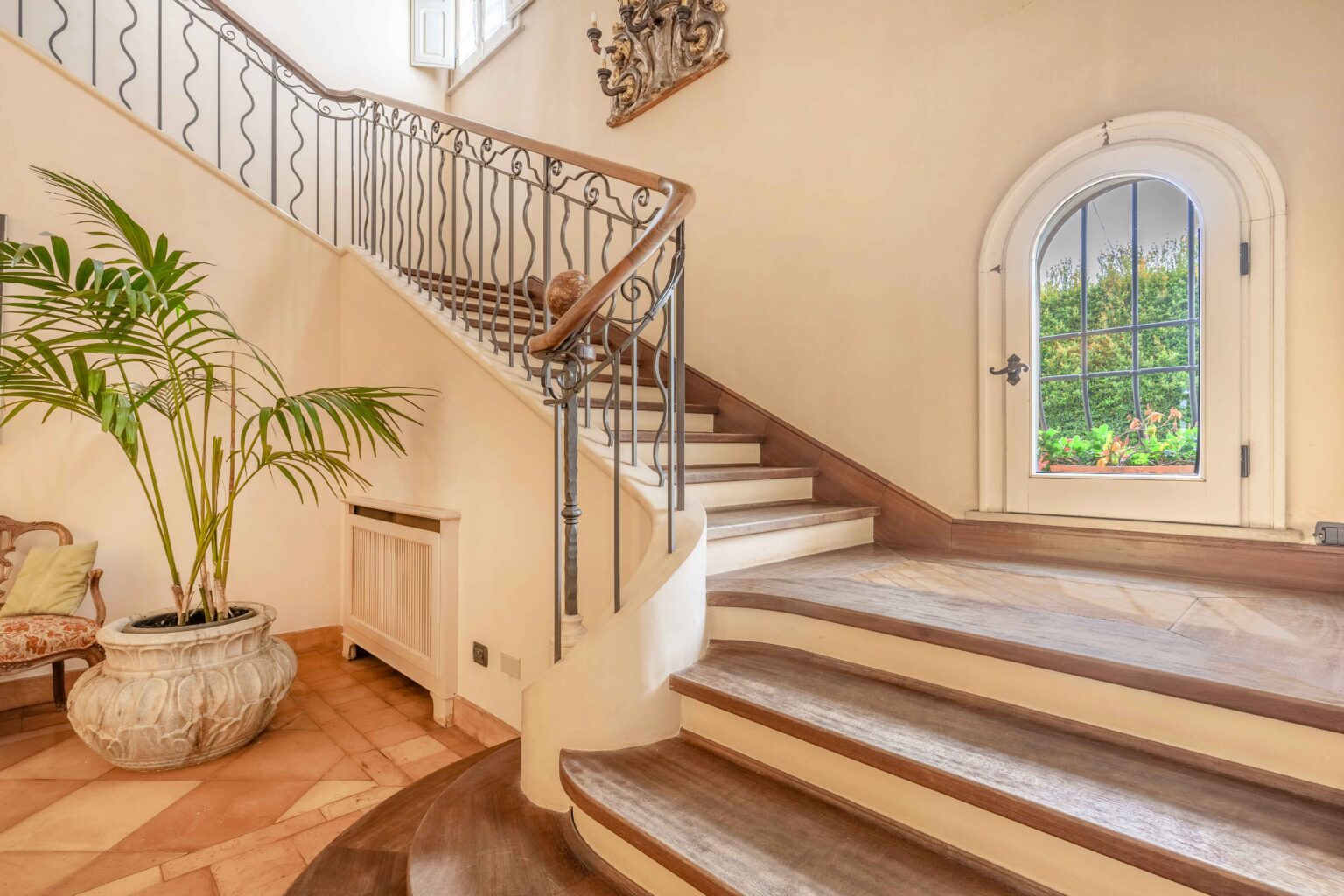 Elegant interior staircase with wooden steps and decorative iron railing, featuring a potted palm plant and arched window that provides natural light and garden views. The warm-toned tiled floor complements the sophisticated design. | Only on SopranoVillas