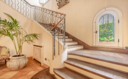 Elegant interior staircase with wooden steps and decorative iron railing, featuring a potted palm plant and arched window that provides natural light and garden views. The warm-toned tiled floor complements the sophisticated design.