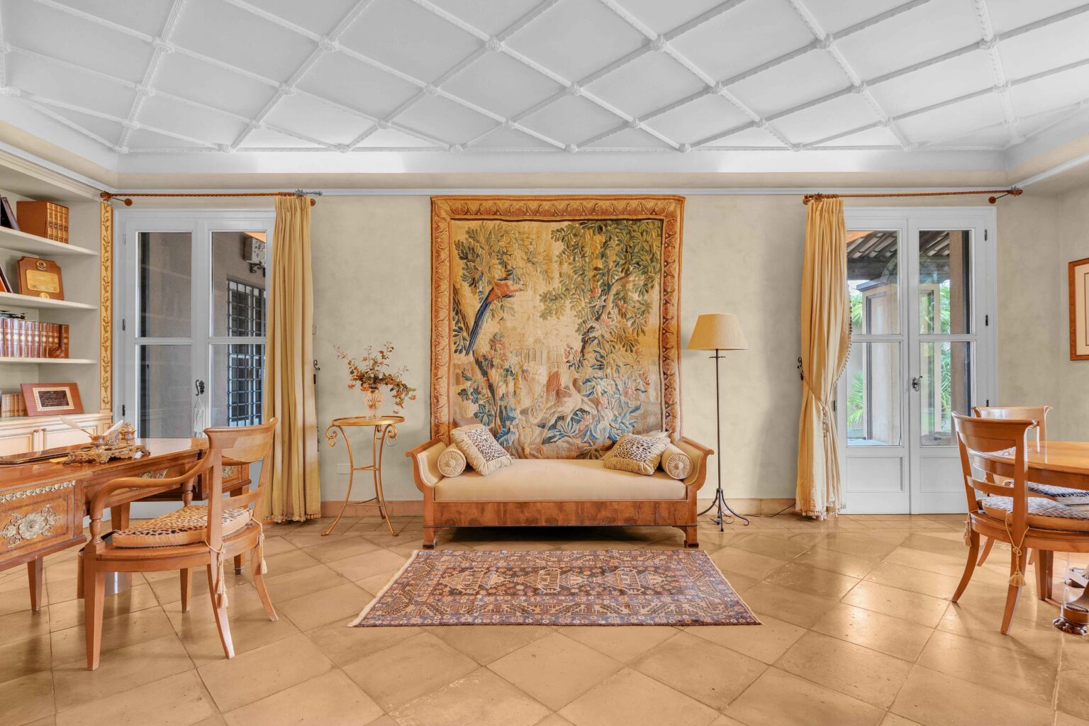 Cozy living room featuring a vintage tapestry on the wall, a beige sofa with decorative pillows, wooden furniture, and a warm color palette. Natural light filters through large windows, enhancing the inviting atmosphere. | Only on SopranoVillas