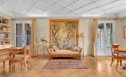 Cozy living room featuring a vintage tapestry on the wall, a beige sofa with decorative pillows, wooden furniture, and a warm color palette. Natural light filters through large windows, enhancing the inviting atmosphere.