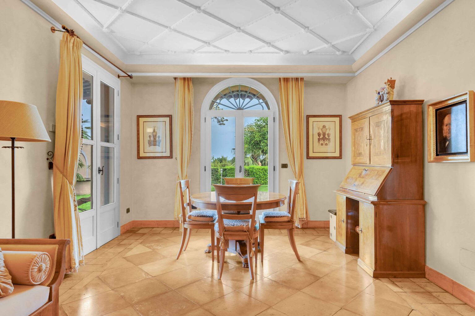 Bright and airy dining area featuring a round wooden table surrounded by four chairs, large windows with yellow curtains, and a wooden cabinet. The room has a light-colored wall and a textured tile floor, creating a warm and inviting atmosphere. | Only on SopranoVillas
