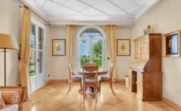 Bright and airy dining area featuring a round wooden table surrounded by four chairs, large windows with yellow curtains, and a wooden cabinet. The room has a light-colored wall and a textured tile floor, creating a warm and inviting atmosphere.