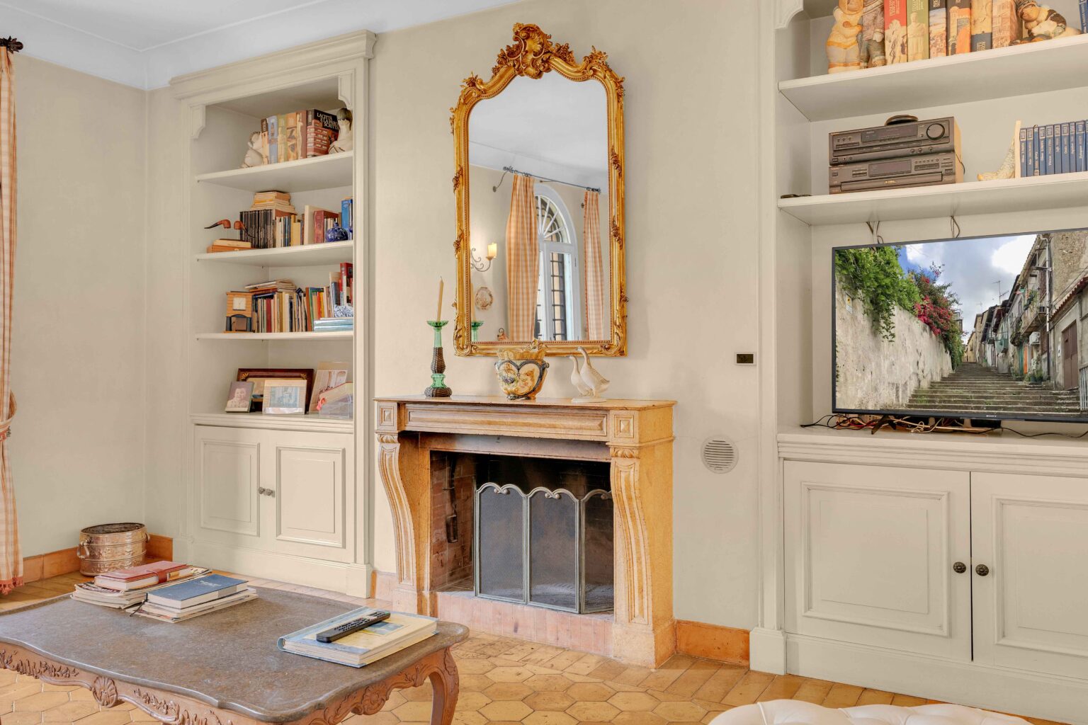 Cozy living room interior featuring a vintage fireplace with ornate detailing, a large golden mirror, bookshelves filled with books, and a television displaying a scenic outdoor view. The space is decorated with warm colors and elegant furnishings, creating an inviting atmosphere. | Only on SopranoVillas
