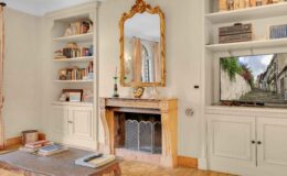 Cozy living room interior featuring a vintage fireplace with ornate detailing, a large golden mirror, bookshelves filled with books, and a television displaying a scenic outdoor view. The space is decorated with warm colors and elegant furnishings, creating an inviting atmosphere.