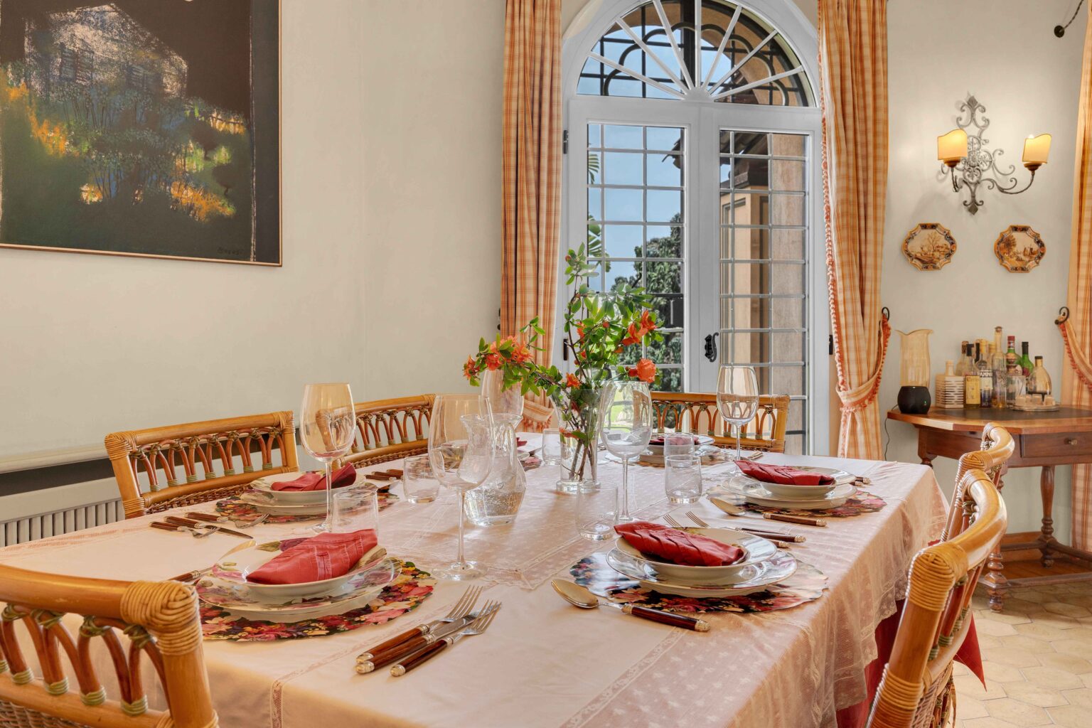 Elegant dining room featuring a beautifully set table with floral placemats, fine china, and crystal glassware. A vase of fresh flowers adds a touch of color, while large windows let in natural light. The decor includes warm tones and artwork, creating a welcoming atmosphere for dining. | Only on SopranoVillas