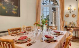 Elegant dining room featuring a beautifully set table with floral placemats, fine china, and crystal glassware. A vase of fresh flowers adds a touch of color, while large windows let in natural light. The decor includes warm tones and artwork, creating a welcoming atmosphere for dining.