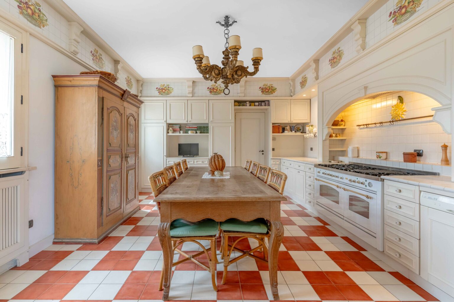 Spacious kitchen featuring a large wooden dining table surrounded by wicker chairs, an antique cabinet, white cabinetry, and a decorative chandelier. The floor displays a classic red and white checkered pattern, while the walls are adorned with floral tiles. Modern appliances blend seamlessly with the traditional design. | Only on SopranoVillas