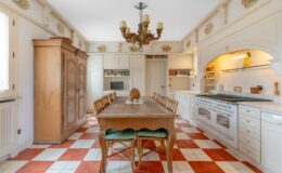 Spacious kitchen featuring a large wooden dining table surrounded by wicker chairs, an antique cabinet, white cabinetry, and a decorative chandelier. The floor displays a classic red and white checkered pattern, while the walls are adorned with floral tiles. Modern appliances blend seamlessly with the traditional design.