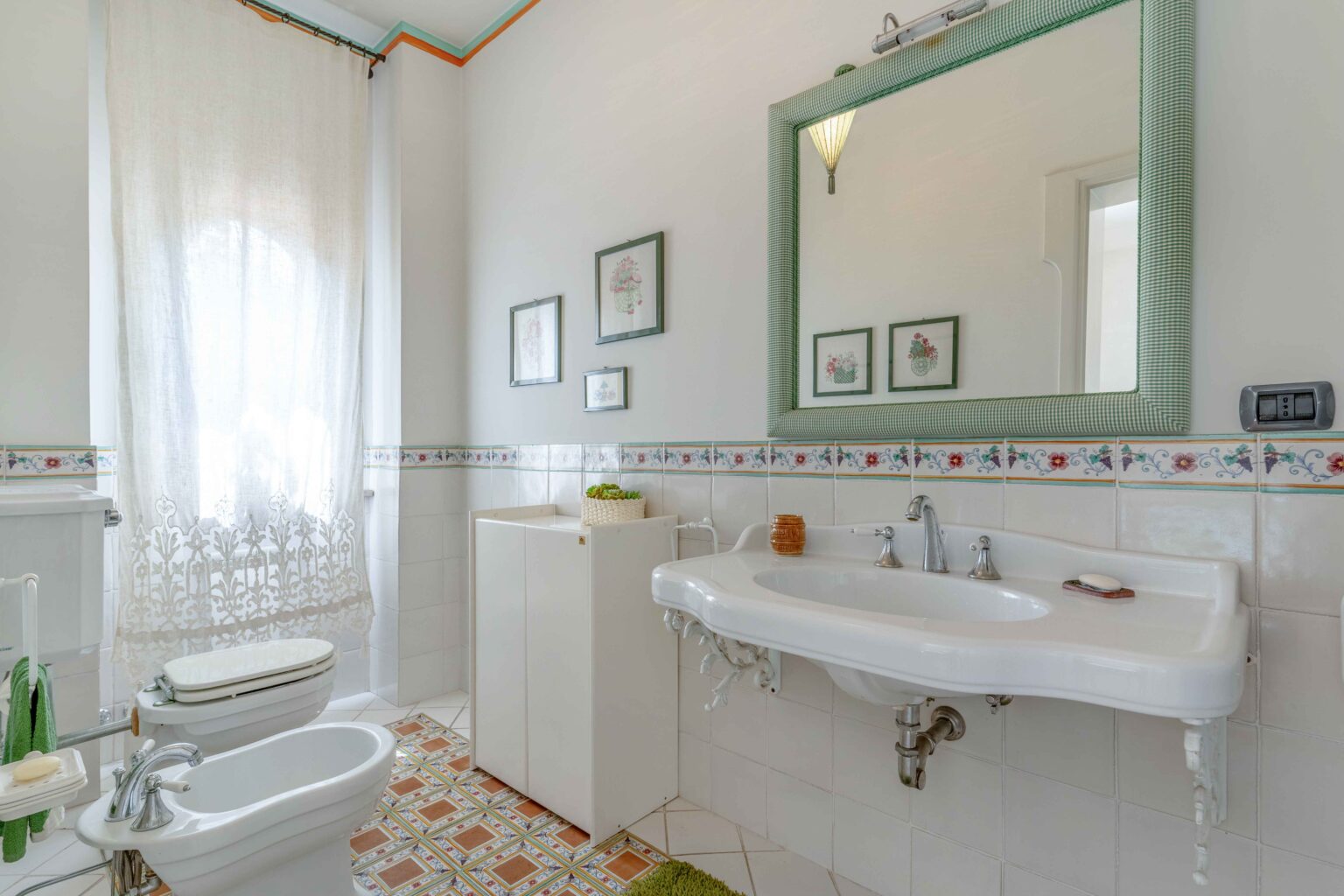 Bright and airy bathroom featuring a white sink, bidet, and toilet, complemented by elegant floral wall decor and a large green-framed mirror. Natural light filters through a sheer curtain, enhancing the modern tile flooring and clean design. | Only on SopranoVillas