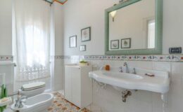 Bright and airy bathroom featuring a white sink, bidet, and toilet, complemented by elegant floral wall decor and a large green-framed mirror. Natural light filters through a sheer curtain, enhancing the modern tile flooring and clean design.