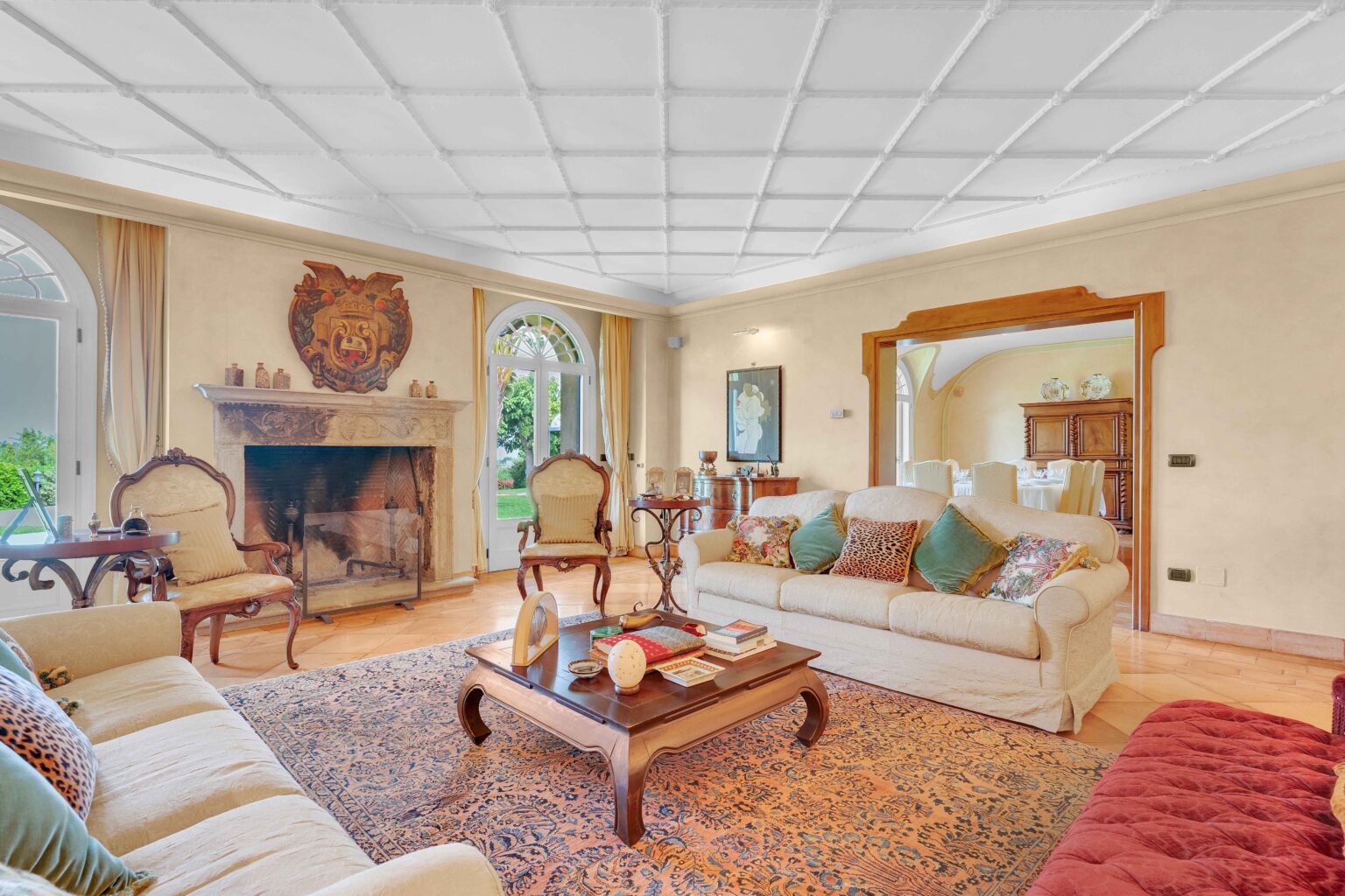 Spacious and elegantly designed living room featuring a cozy fireplace, plush sofas with decorative cushions, and a wooden coffee table adorned with books and decor. The room has large windows that provide natural light and a view of the garden, complemented by a beautiful area rug and classic furniture. | Only on SopranoVillas