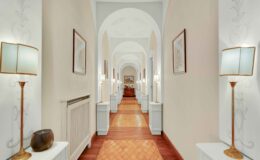 Elegant hotel corridor featuring arched ceilings, decorative artwork, and stylish lamps. The warm wooden flooring complements the light-colored walls, creating a welcoming atmosphere.
