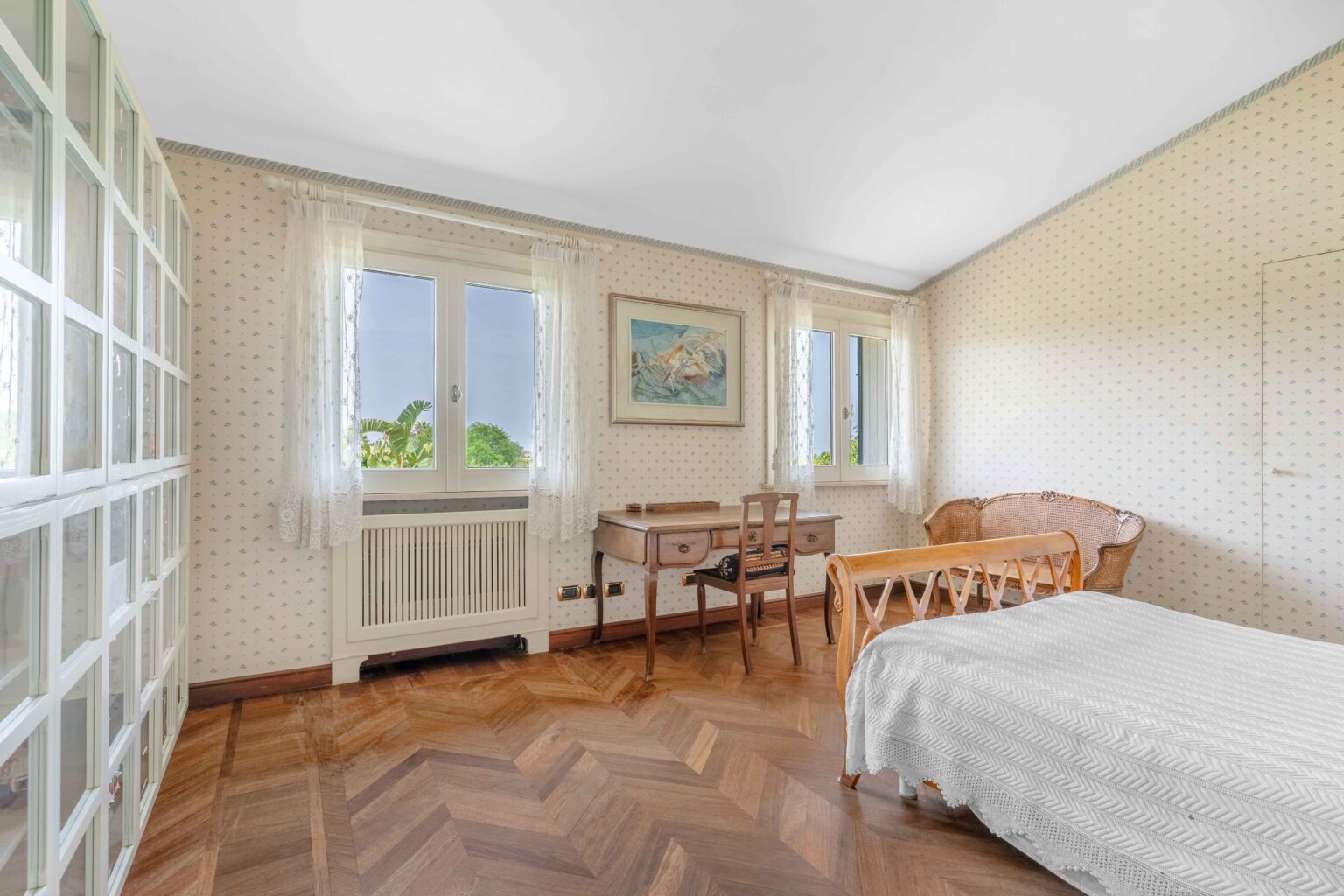 Bright and airy bedroom featuring a wooden floor, a white bedspread, a vintage desk with a chair, and large windows adorned with lace curtains. The room has floral wallpaper and a cozy wicker chair, creating a warm and inviting atmosphere. | Only on SopranoVillas