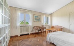 Bright and airy bedroom featuring a wooden floor, a white bedspread, a vintage desk with a chair, and large windows adorned with lace curtains. The room has floral wallpaper and a cozy wicker chair, creating a warm and inviting atmosphere.