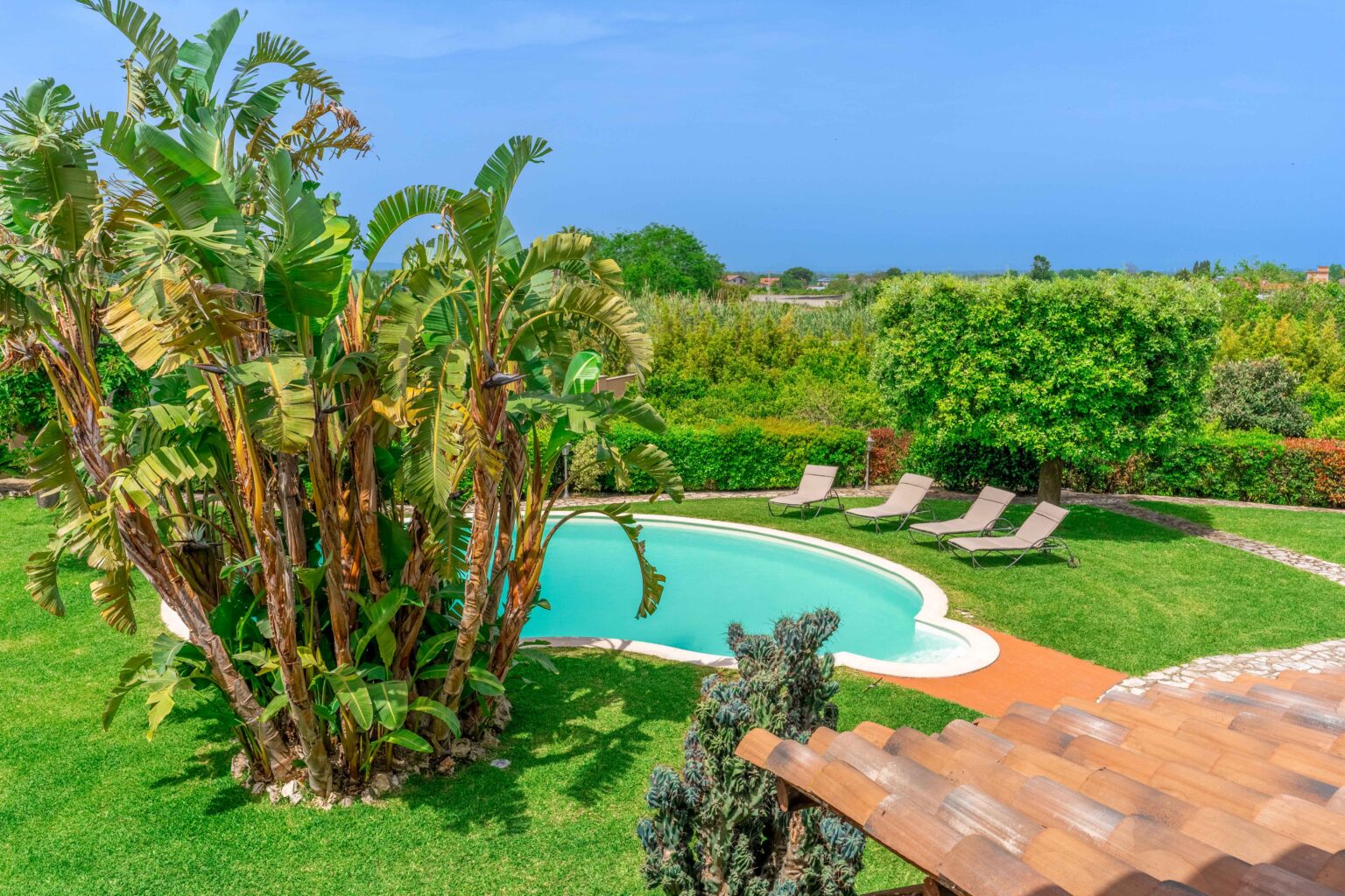 Lush tropical garden featuring a swimming pool surrounded by lounge chairs, with vibrant greenery and a clear blue sky in the background. Ideal for relaxing and enjoying outdoor leisure activities. | Only on SopranoVillas
