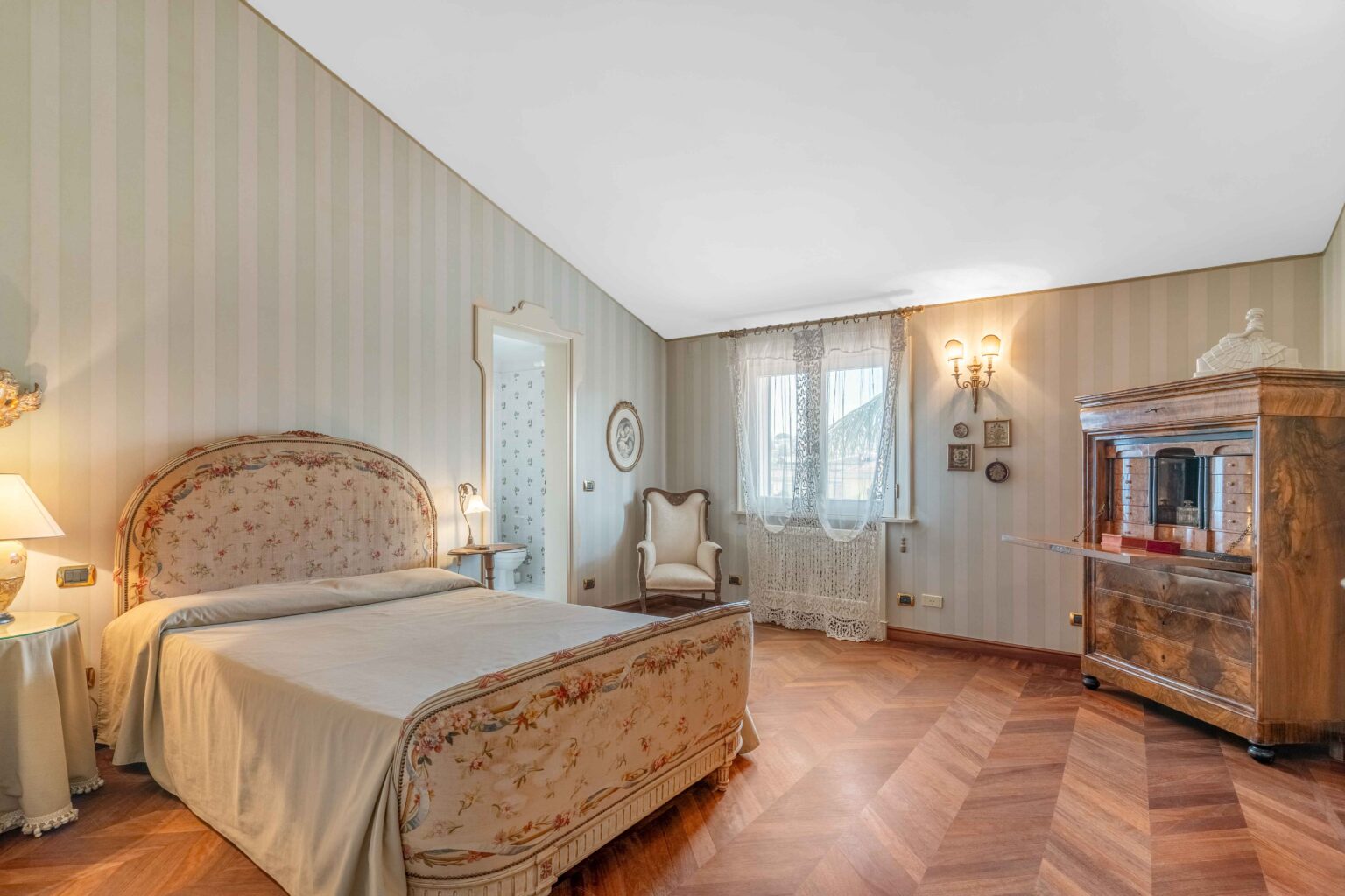 Elegant bedroom featuring a vintage floral-patterned headboard, wooden flooring, and soft lighting. The room includes a cozy armchair, a decorative wooden cabinet, and lace curtains framing the window, creating a warm and inviting atmosphere. | Only on SopranoVillas
