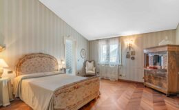 Elegant bedroom featuring a vintage floral-patterned headboard, wooden flooring, and soft lighting. The room includes a cozy armchair, a decorative wooden cabinet, and lace curtains framing the window, creating a warm and inviting atmosphere.