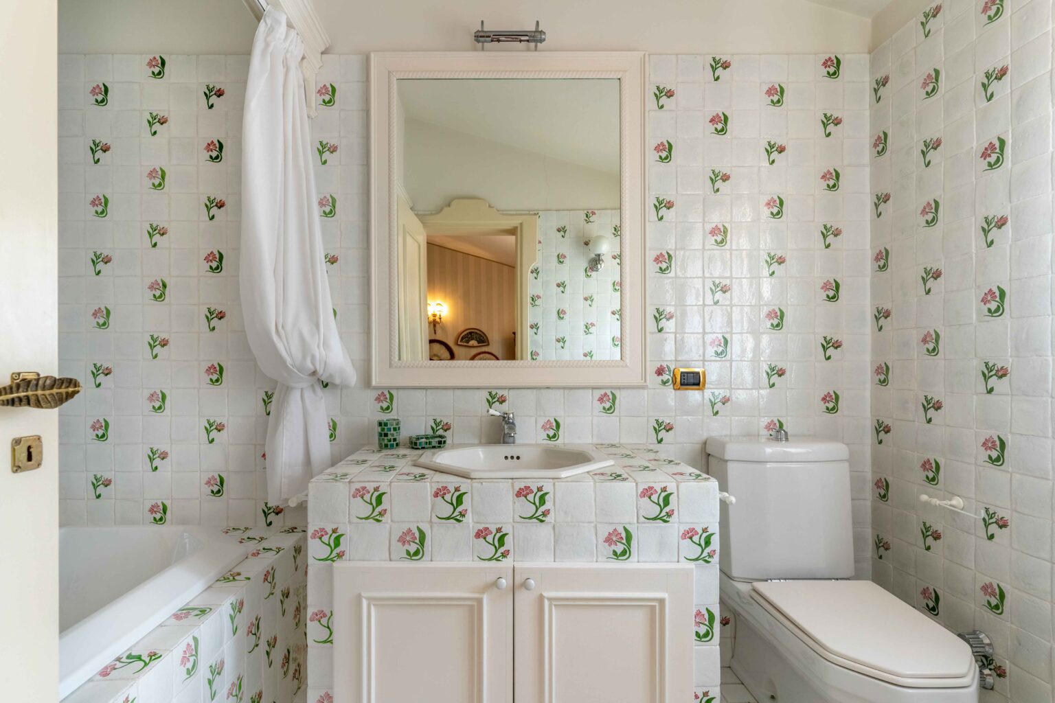 Bright and charming bathroom featuring floral-patterned tiles, a white bathtub, a modern toilet, and a stylish sink with a mirror. The decor combines classic and contemporary elements, creating an inviting atmosphere. | Only on SopranoVillas