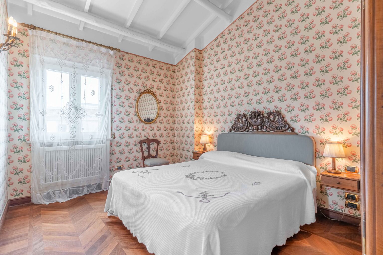 Cozy vintage bedroom featuring floral wallpaper, a decorative mirror, and a wooden chair. The room includes a plush bed with a white coverlet and elegant lighting, creating a warm and inviting atmosphere. | Only on SopranoVillas
